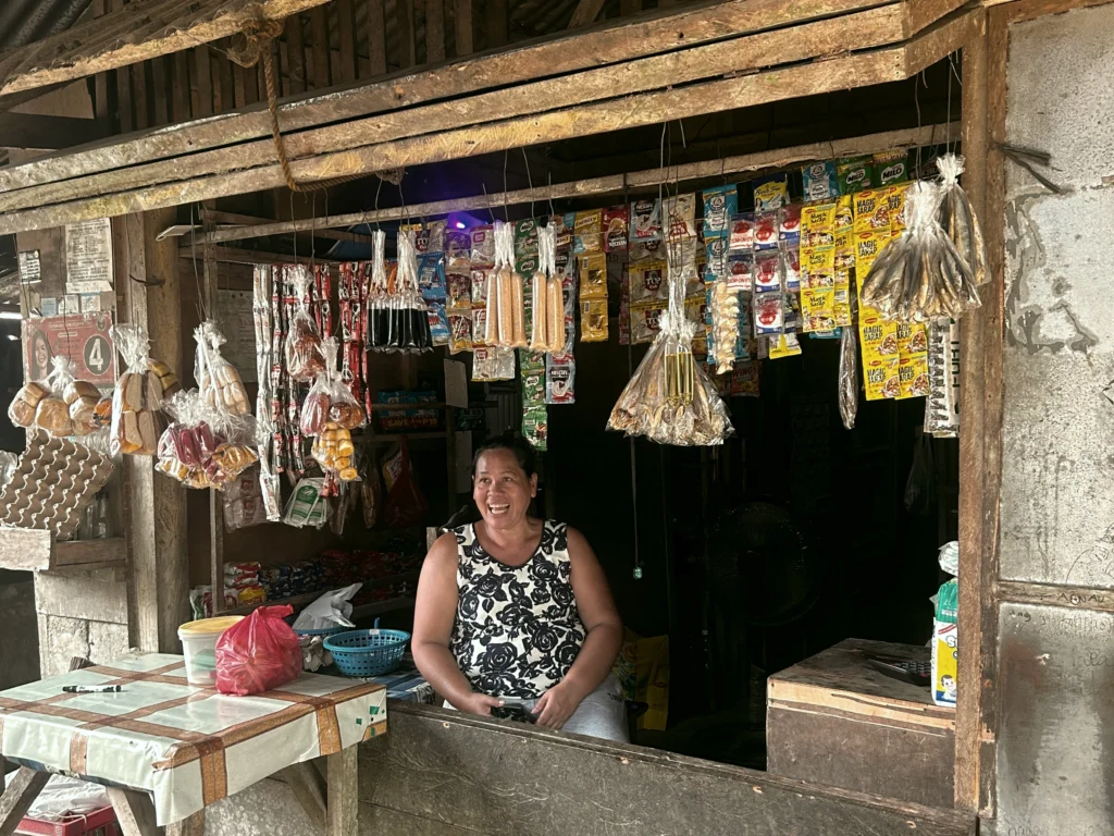 A cheerful local Filipino woman smiling warmly from inside her small neighborhood shop, welcoming visitors during a community mission trip to the Philippines.