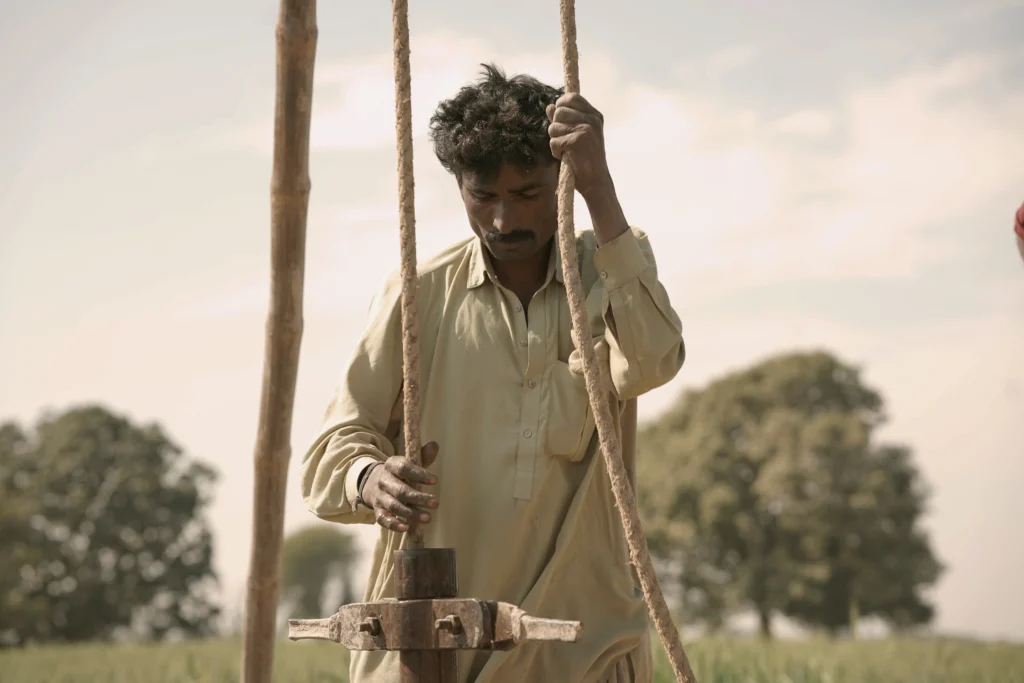 A close-up shot of a man wearing a light-colored shirt, carefully holding onto ropes to operate traditional well-digging machinery in an open, grassy field under a bright sky.