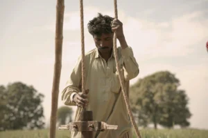 A close-up shot of a man wearing a light-colored shirt, carefully holding onto ropes to operate traditional well-digging machinery in an open, grassy field under a bright sky.