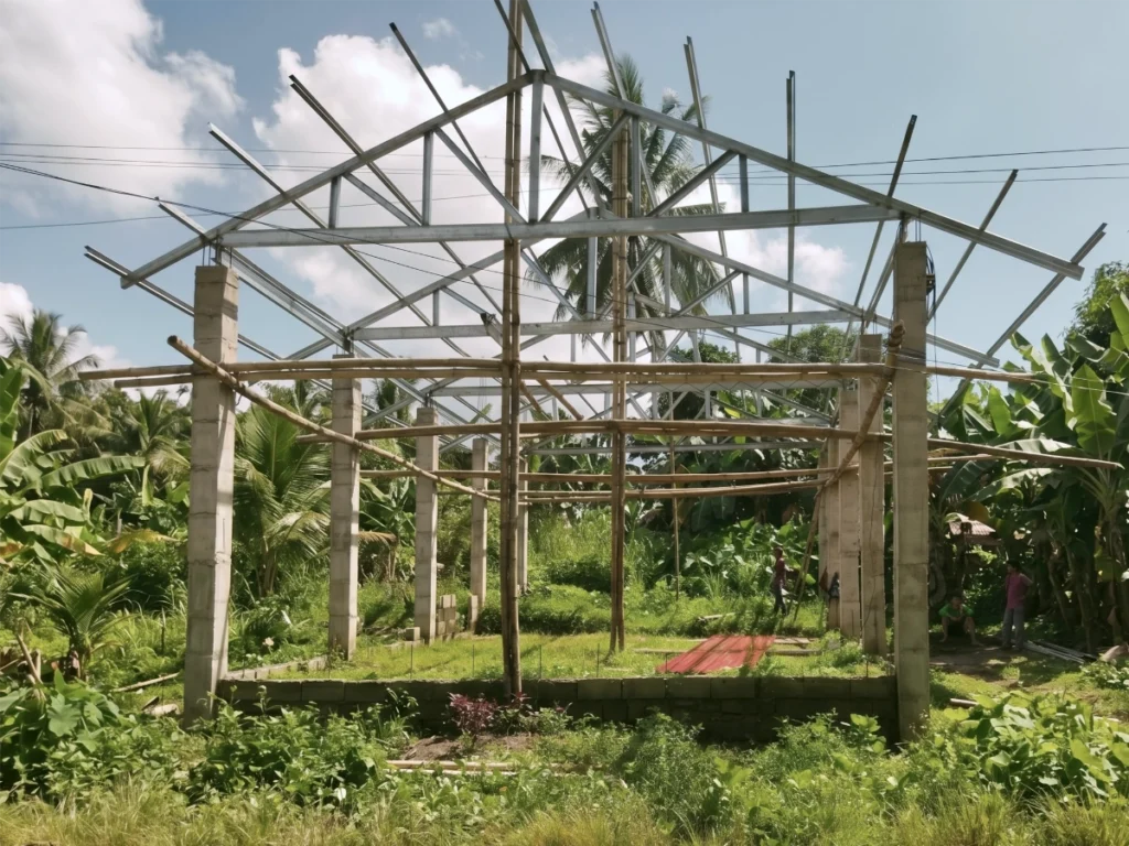 The structural metal roof trusses and concrete pillars of an open-air church building under construction in a tropical region.