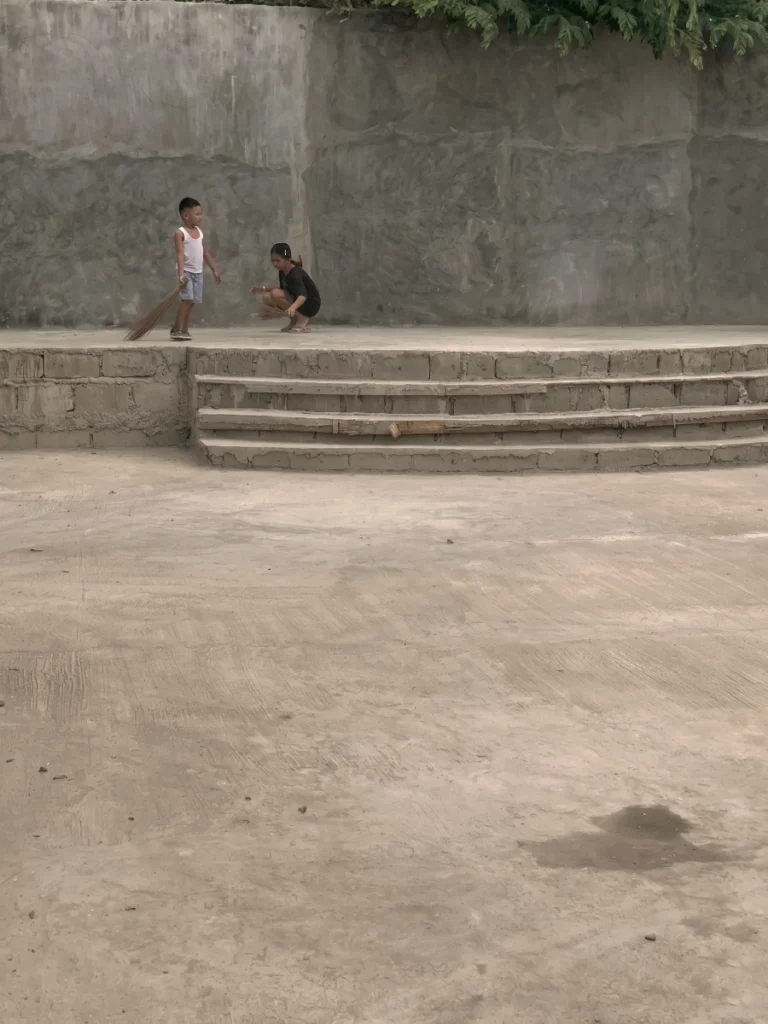 A wide-angle shot of a young boy and a girl crouching to sweep a large, empty concrete stage with tiered steps. The background features a tall, plain concrete wall, emphasizing the scale of the ongoing construction project.
