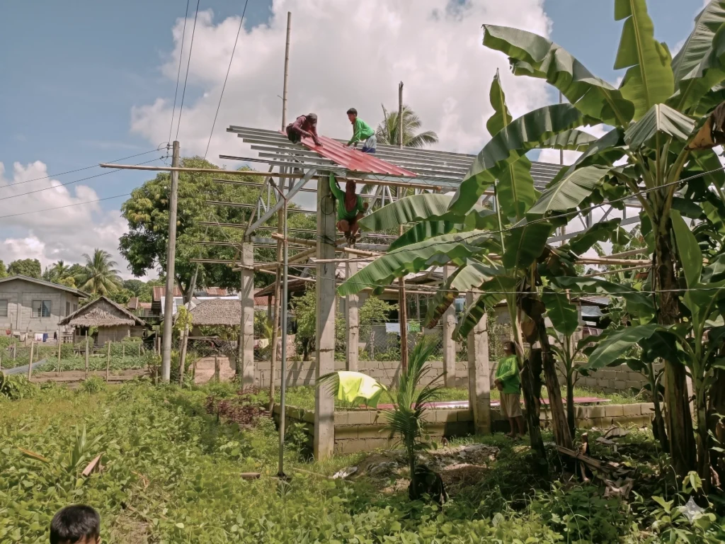 Construction crew installing red corrugated roofing panels on a large open-air church building frame surrounded by tropical trees.