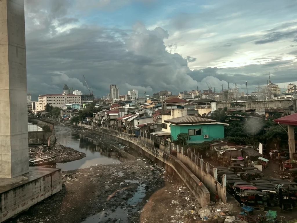 A view of a Philippine city showing a polluted canal lined with densely packed informal housing, contrasting sharply with modern skyscrapers and construction cranes in the distance.
