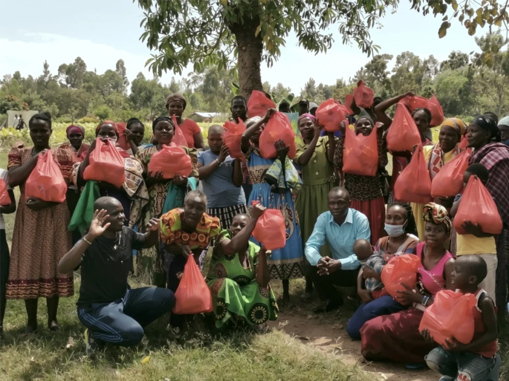 A large group of local villagers gathered under a tree holding up red packages of essential food items and supplies.