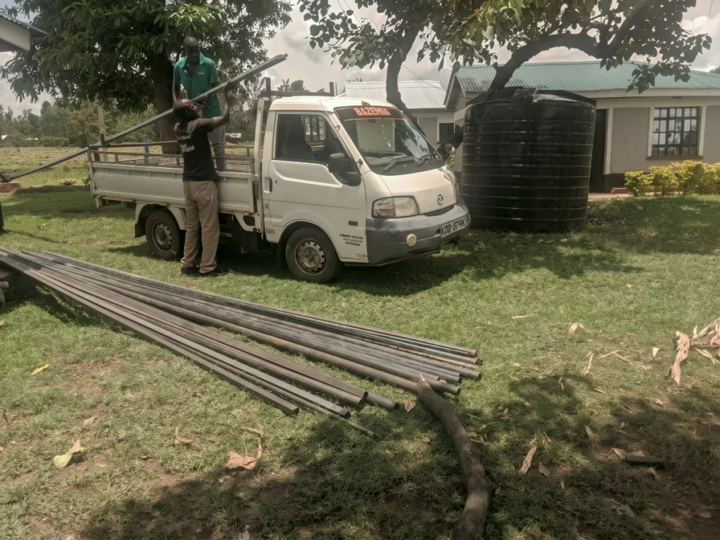 Two workers unload long steel beams from a small white truck next to a large black water tank, gathering materials to build a clean water tower for a community in Kenya.