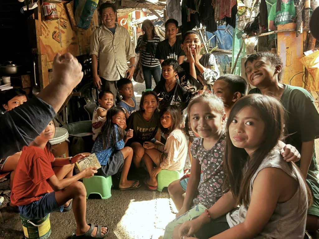 A large group of happy Filipino children and adults smiling, laughing, and looking warmly toward the camera while gathered together in a rustic neighborhood setting.