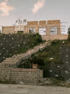 A wide-angle shot of a building under construction atop a grassy hillside. The structure is made of concrete blocks, with a sign reading "King Jesus Christian Fellowship." A stone staircase leads up the slope towards the building, which is situated above a steep embankment covered in protective netting.