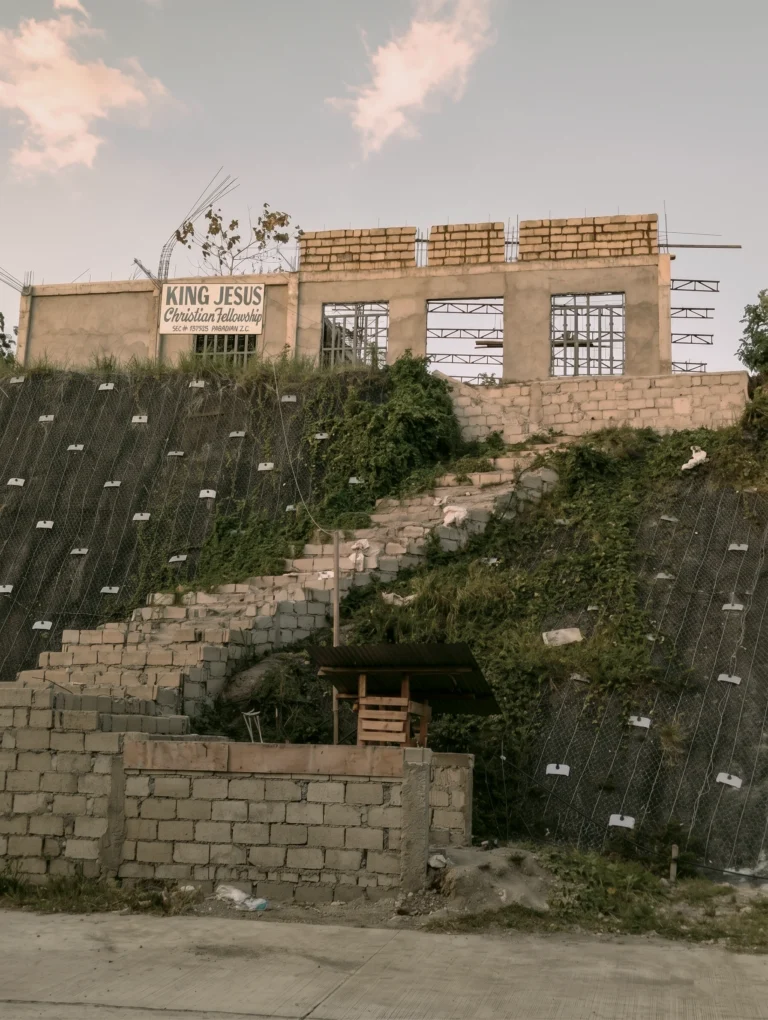A wide-angle shot of a building under construction atop a grassy hillside. The structure is made of concrete blocks, with a sign reading "King Jesus Christian Fellowship." A stone staircase leads up the slope towards the building, which is situated above a steep embankment covered in protective netting.