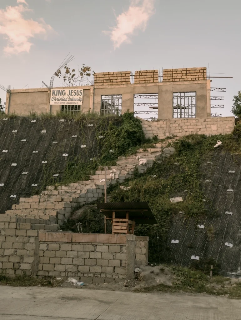 A wide-angle shot of a building under construction atop a grassy hillside. The structure is made of concrete blocks, with a sign reading "King Jesus Christian Fellowship." A stone staircase leads up the slope towards the building, which is situated above a steep embankment covered in protective netting.