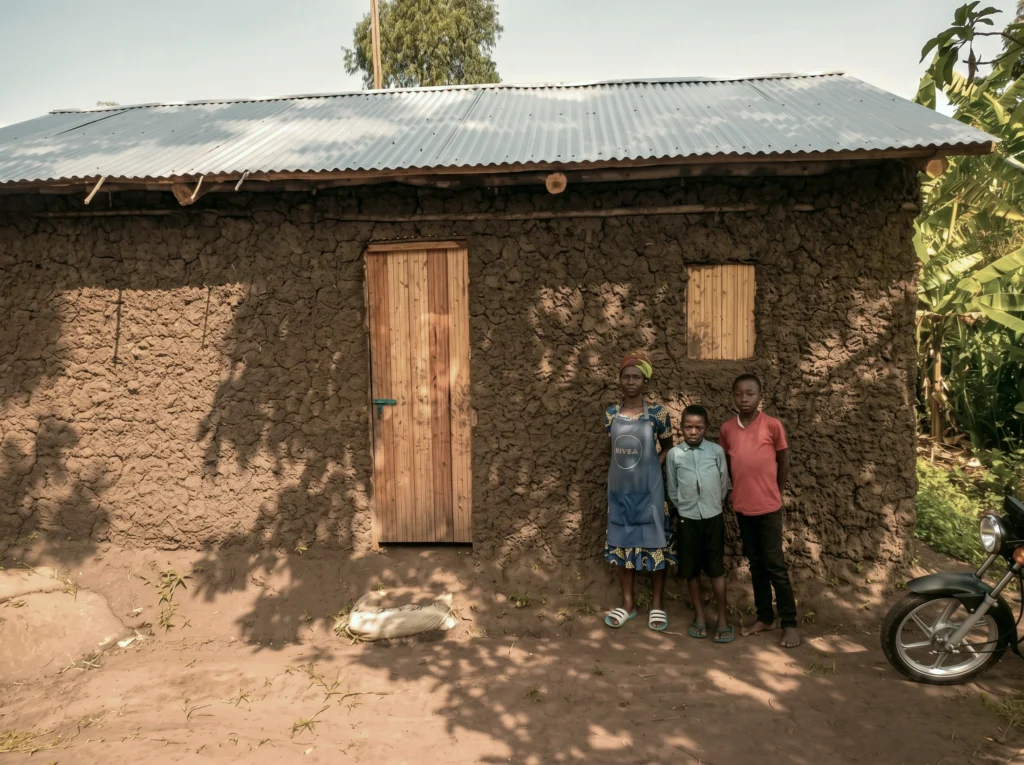 A mother and her two sons stand together in front of their newly constructed mud-wall home with a secure metal roof, marking their family's transition from ruin to restoration.