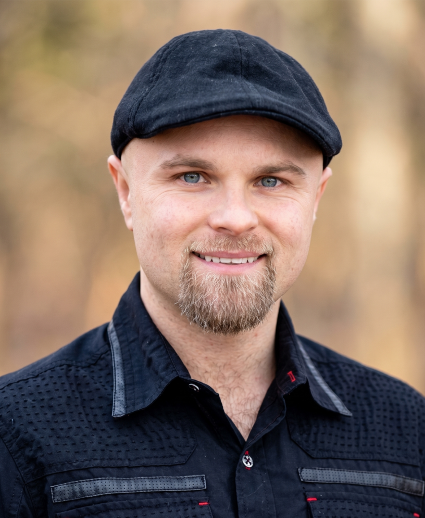 A portrait photo of a smiling man with a goatee, wearing a black flat cap and a textured black collared shirt, outdoors with a natural, blurred background.
