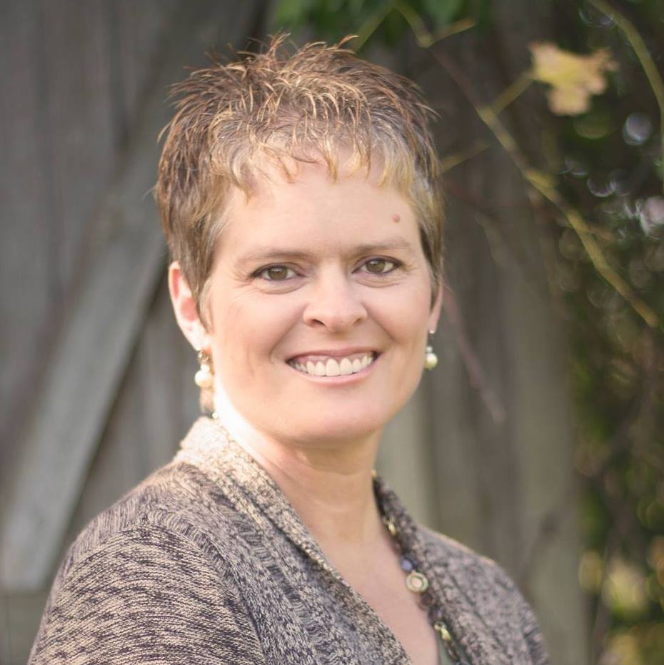 A warm, professional headshot of Lisa Beth Adams smiling, with short hair and wearing a textured sweater, set against a soft, natural background.
