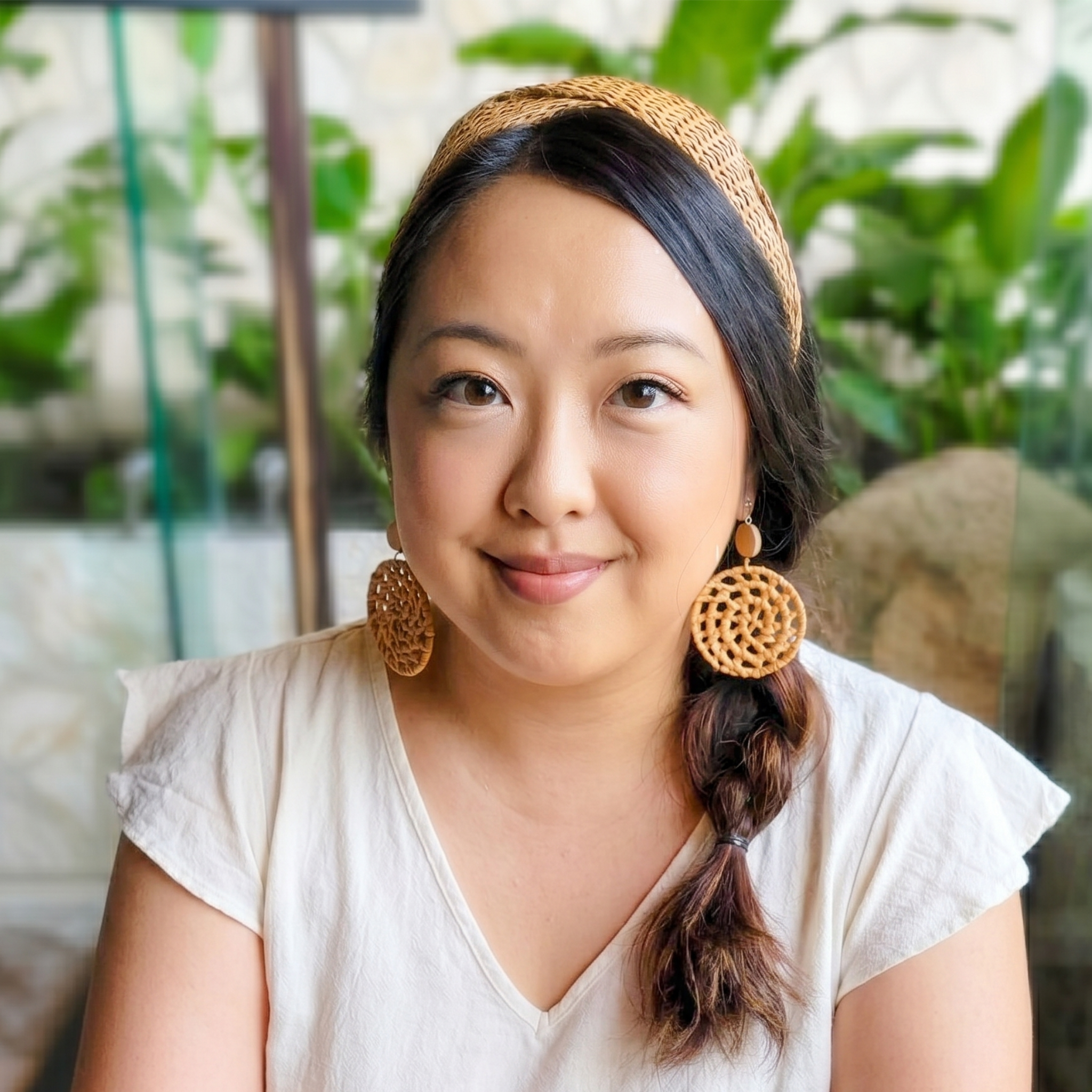 A portrait of a woman with dark hair styled in a side braid, wearing a woven headband and round, textured tan earrings, smiling while facing the camera with a soft-focus plant background.