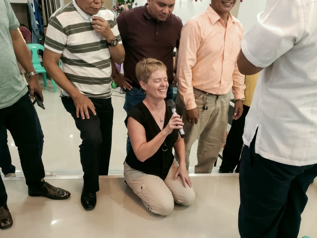 Missionary Lisa Beth Adams kneeling on the floor with a microphone, laughing joyfully with her eyes closed while surrounded by smiling local church leaders during a Philippine mission service.