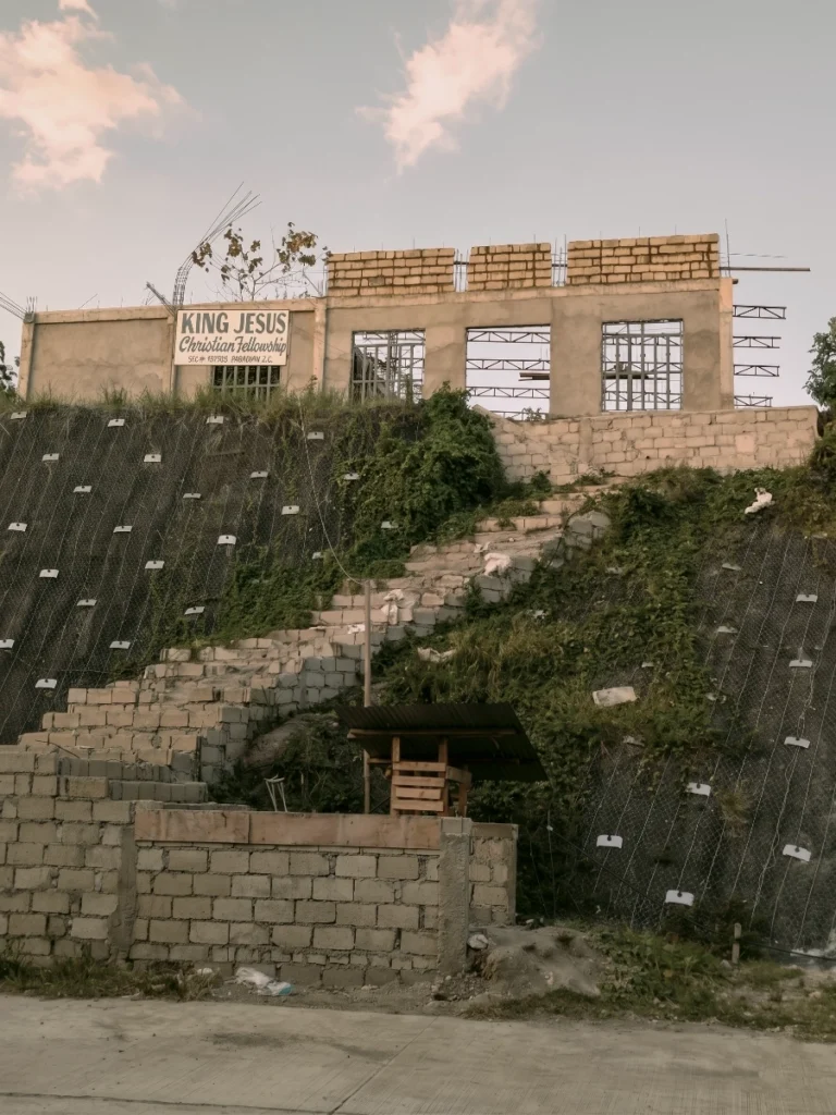 A wide-angle shot of a building under construction atop a grassy hillside. The structure is made of concrete blocks, with a sign reading "King Jesus Christian Fellowship." A stone staircase leads up the slope towards the building, which is situated above a steep embankment covered in protective netting.