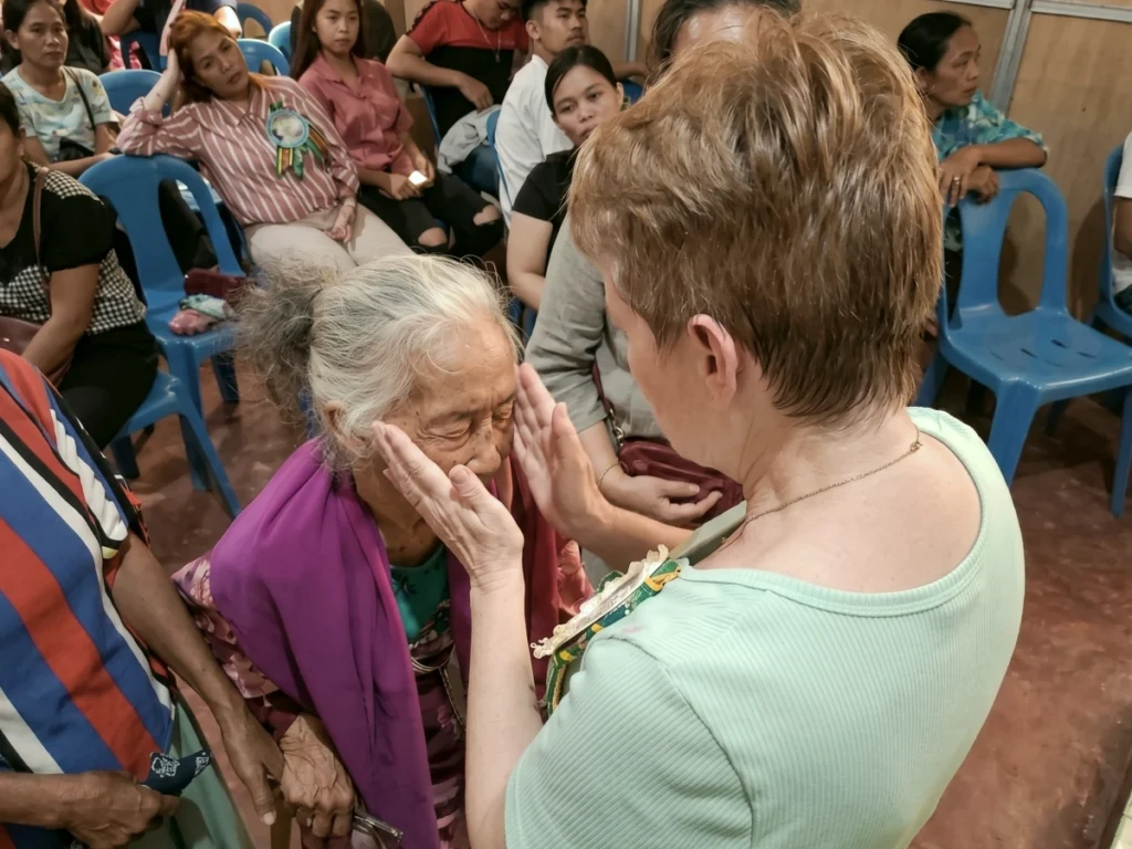 Missionary Lisa Beth Adams gently holding her hands near an elderly Filipino woman's face, engaging in deep, compassionate prayer during a community church service.