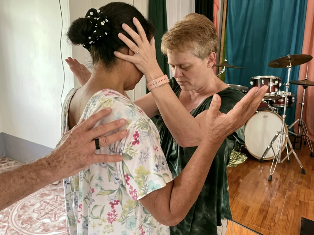 Missionary Lisa Beth Adams gently placing her hands on a local Filipino woman's head, eyes closed in deep, faithful prayer during a heartfelt church service.