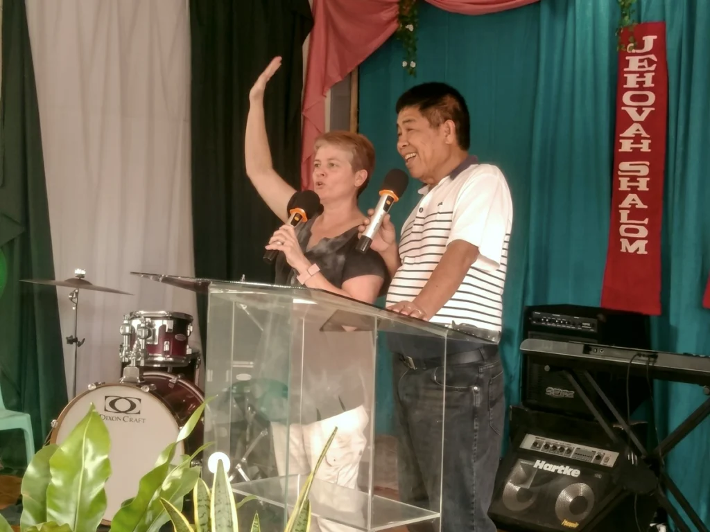 Visiting missionary Lisa Beth Adams stands at a clear podium with her hand raised, passionately preaching a sermon with a microphone alongside a smiling local pastor during a mission worship service.
