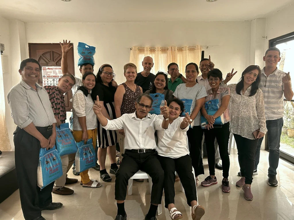 A diverse group of missionaries and local church leaders posing for a happy group photo, holding bags of premium rice during a community fellowship meeting in the Philippines.