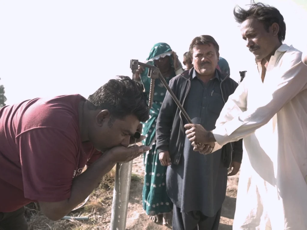 A man in a maroon shirt drinks water directly from a metal pipe while another man operates the pump; a woman and another man stand in the background, witnessing the completion of the life-saving infrastructure.