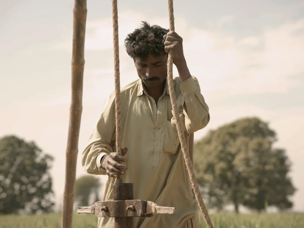 A close-up shot of a man wearing a light-colored shirt, carefully holding onto ropes to operate traditional well-digging machinery in an open, grassy field under a bright sky.