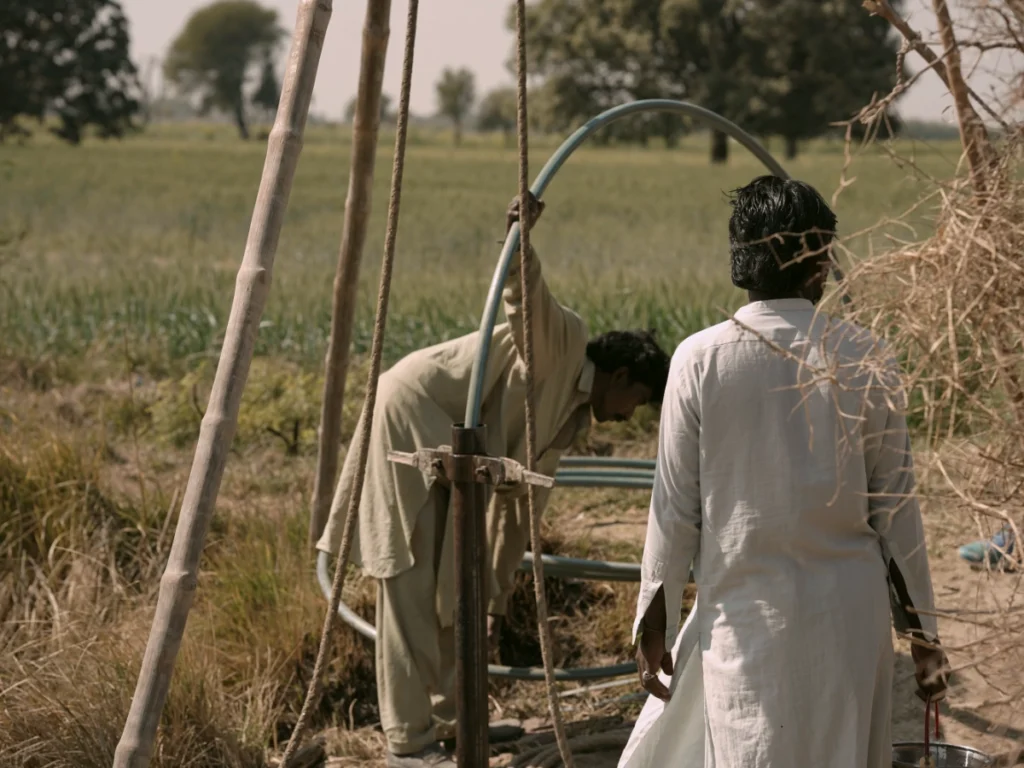 Two men in traditional light-colored clothing work on installing a curved pipe into a well-digging site in a green field; one man is bending over the machinery while the other stands nearby.