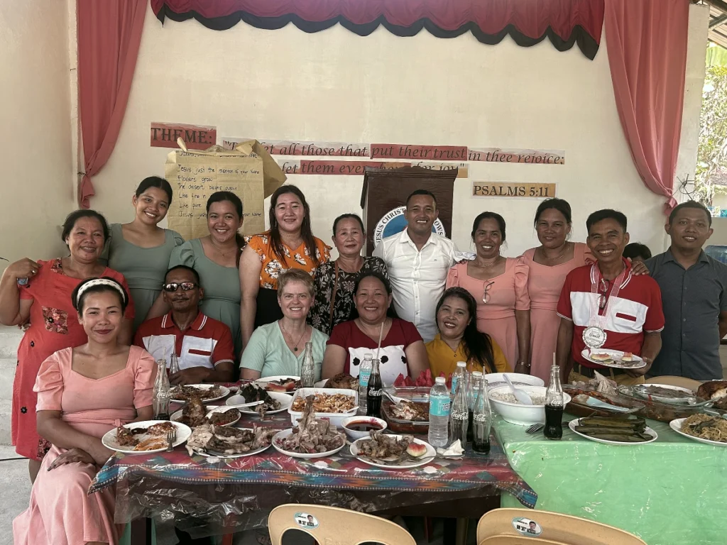A joyful group of Philippine church members and visiting mission partners smiling together behind a long banquet table filled with a fellowship feast, celebrating their new family bond.