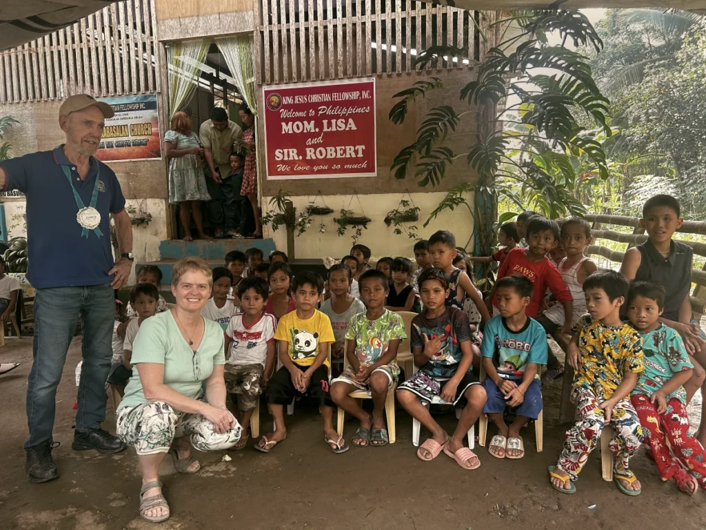 Missionaries Lisa Beth Adams and Robert Hagen smiling warmly while posing with a large group of local Filipino children seated in front of a church building featuring a red welcome banner.