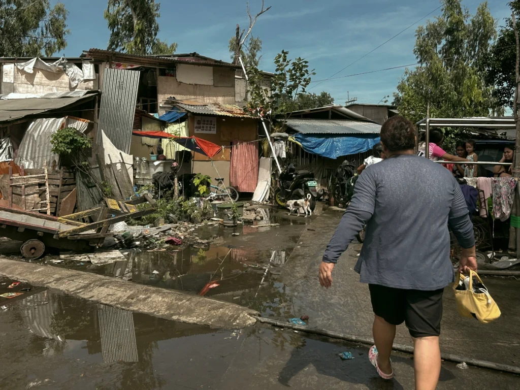 The back of a person walking through large puddles of stagnant water on a rough road, surrounded by informal homes built from scrap wood, corrugated metal, and tarps.