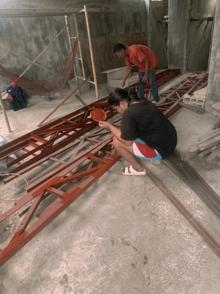 A woman crouching while painting a reddish-brown primer onto a large steel truss, with a man standing in the background also painting another section of the metal framework inside a workspace.