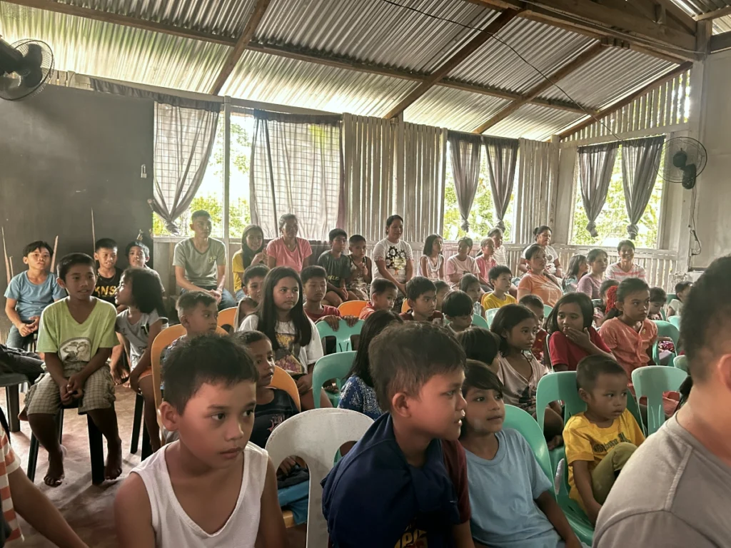 A large group of local Filipino children and parents sitting attentively in colorful plastic chairs inside a rustic community hall during a mission outreach gathering.