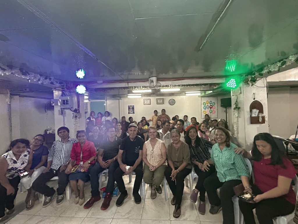A large, welcoming group of local church members and visiting mission partners sitting together in rows, smiling for a group photo during a community fellowship gathering in the Philippines.