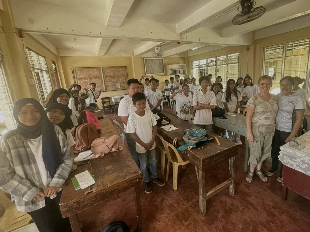 A visiting mission partner smiles warmly while posing with a large, joyful group of local Filipino students inside their classroom during a community outreach trip.