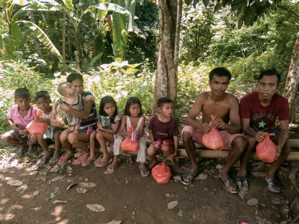 A group of nine people, including adults and young children, sitting on a bamboo pole in a tropical forest environment, holding orange bags of emergency relief supplies.