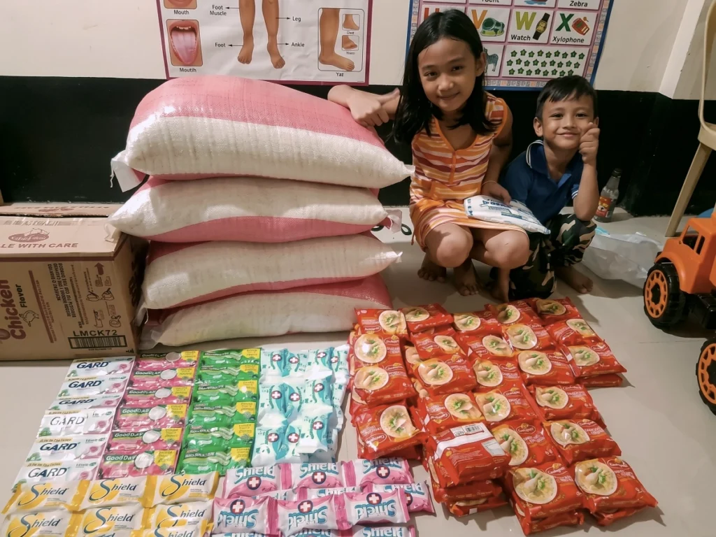 Two smiling children crouching next to large sacks of rice and organized rows of dry goods, preparing to pack and distribute relief supplies to flood victims in the Philippines.