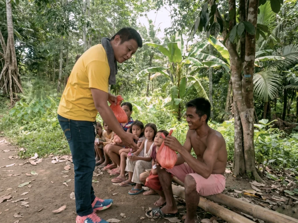 A man in a yellow shirt hands emergency relief bags to a father and his children sitting on a bamboo log in a rural Philippine mountain village recovering from torrential flooding.