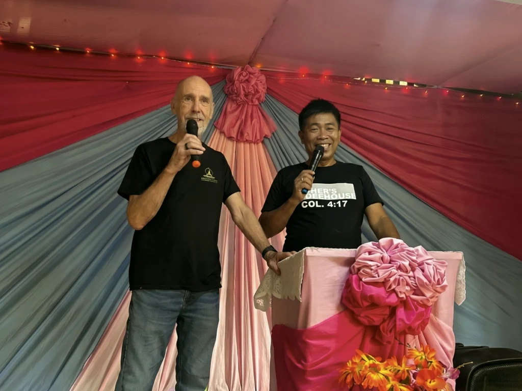 Visiting missionary Pastor Robert Hagen and a local Filipino pastor smiling joyfully while co-preaching a sermon together with microphones during a mission trip church service.