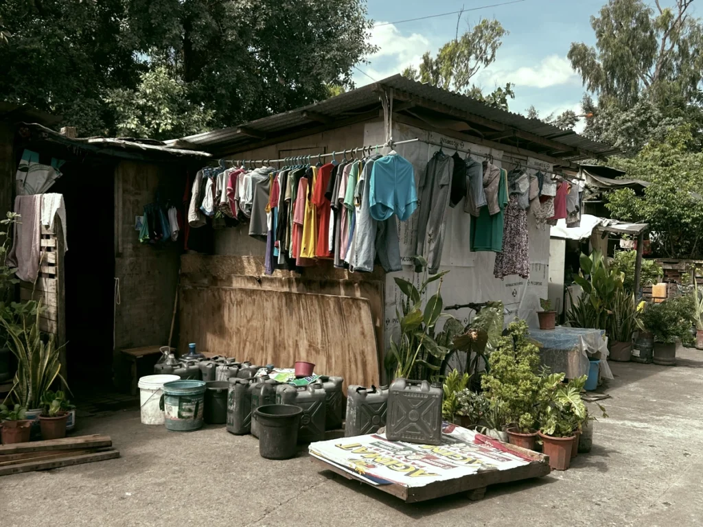 A humble neighborhood scene in the Philippines featuring a small residential structure with a corrugated roof, a long line of drying clothes, and numerous dark plastic water containers resting on the concrete ground.