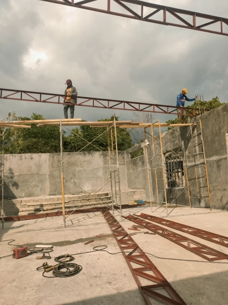 A construction site scene showing two men working high up on metal scaffolding to install long, red-painted steel roof trusses onto the concrete block walls of a building. Several other unassembled trusses lie on the concrete floor in the foreground.