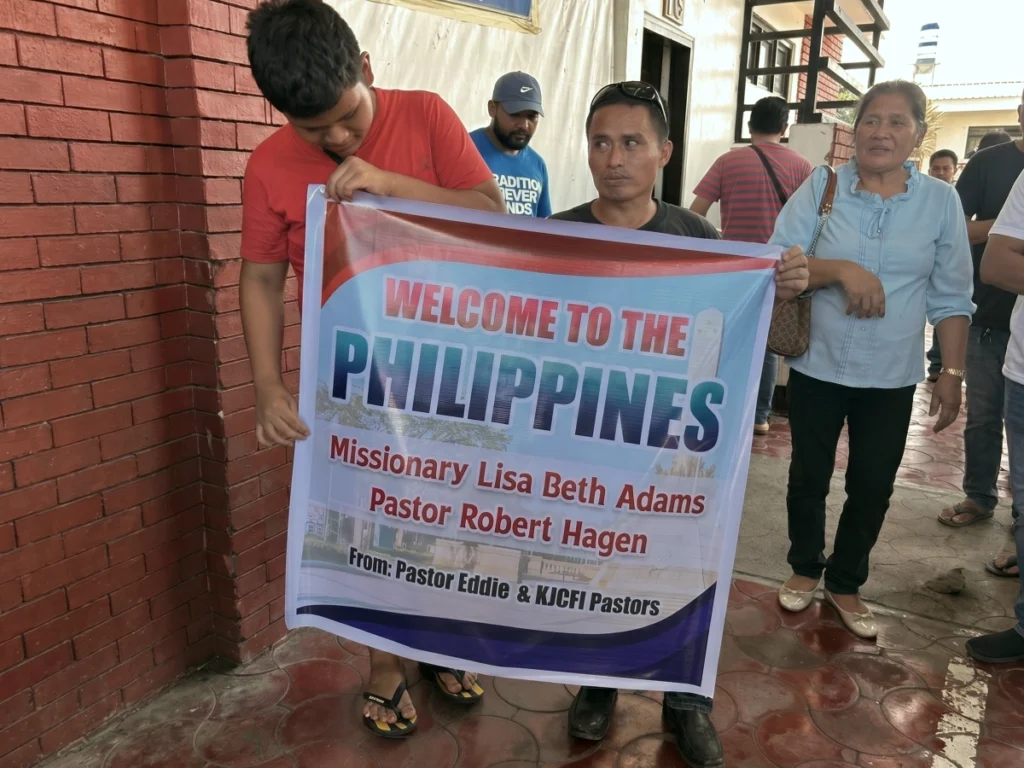 Local church members holding a large printed banner reading "Welcome to the Philippines" to warmly greet arriving missionaries Lisa Beth Adams and Pastor Robert Hagen.
