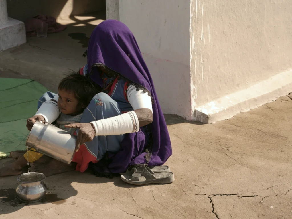 A woman wearing a purple veil and white bracelets sits on the dry earth next to a white wall, pouring water from a metal pitcher into a small bowl held by a young child sitting in her lap.