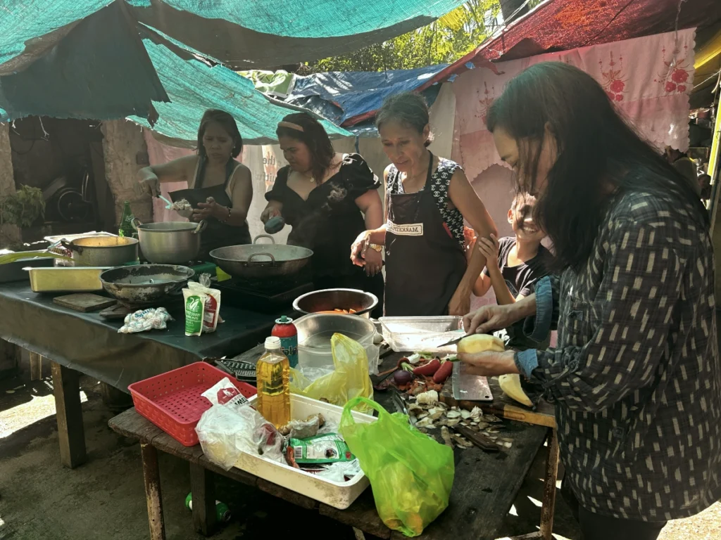 A group of local Filipino women working joyfully together in an outdoor kitchen, chopping vegetables and stirring pots to prepare a large fellowship meal for the mission gathering.