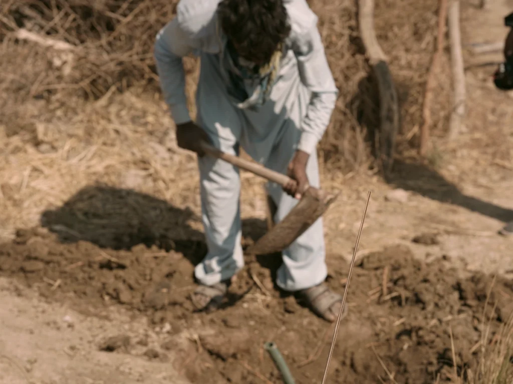 A worker wearing light-colored traditional clothing uses a shovel to dig a trench in dry, rural soil as part of a mission project to bring clean drinking water to a remote area in Pakistan.