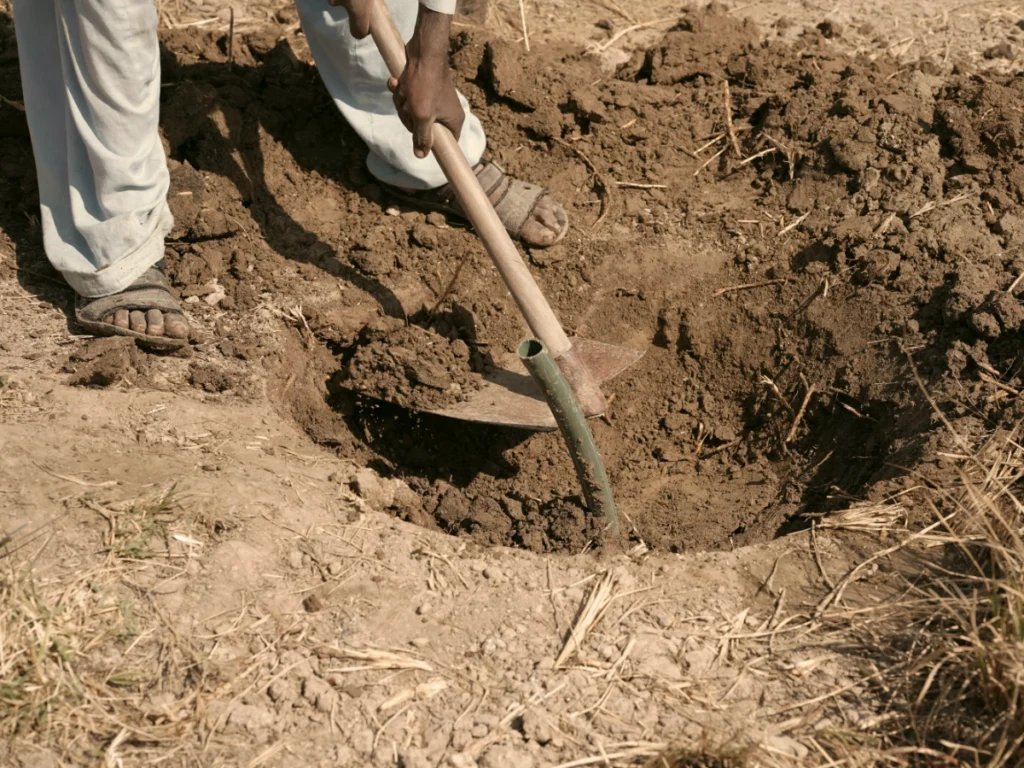 An overhead close-up shot of a person's hands using a metal shovel to dig soil away from a section of green pipe, showing the physical labor necessary to establish a foundation for clean water access.