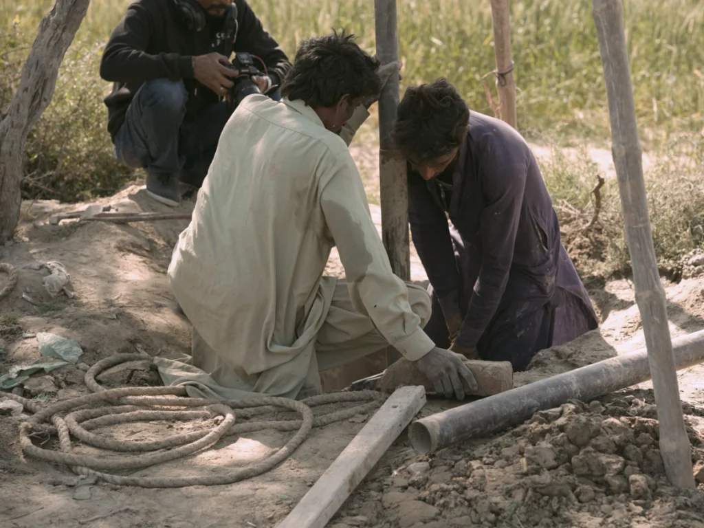 A close-up shot of two men in traditional clothing kneeling in the dirt to work on a pipe installation for a new well, with a photographer crouched in the background capturing the construction process.