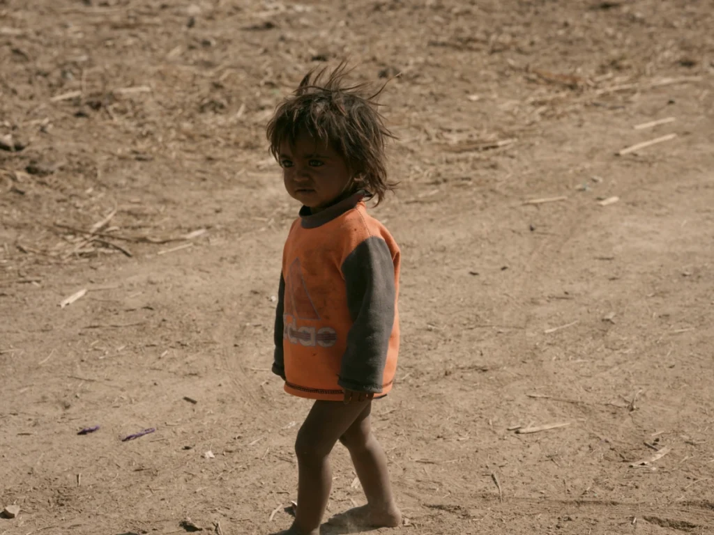 A full-body shot of a young child wearing an orange and gray long-sleeved shirt walking across a barren, dirt-covered ground in a rural area of Pakistan.