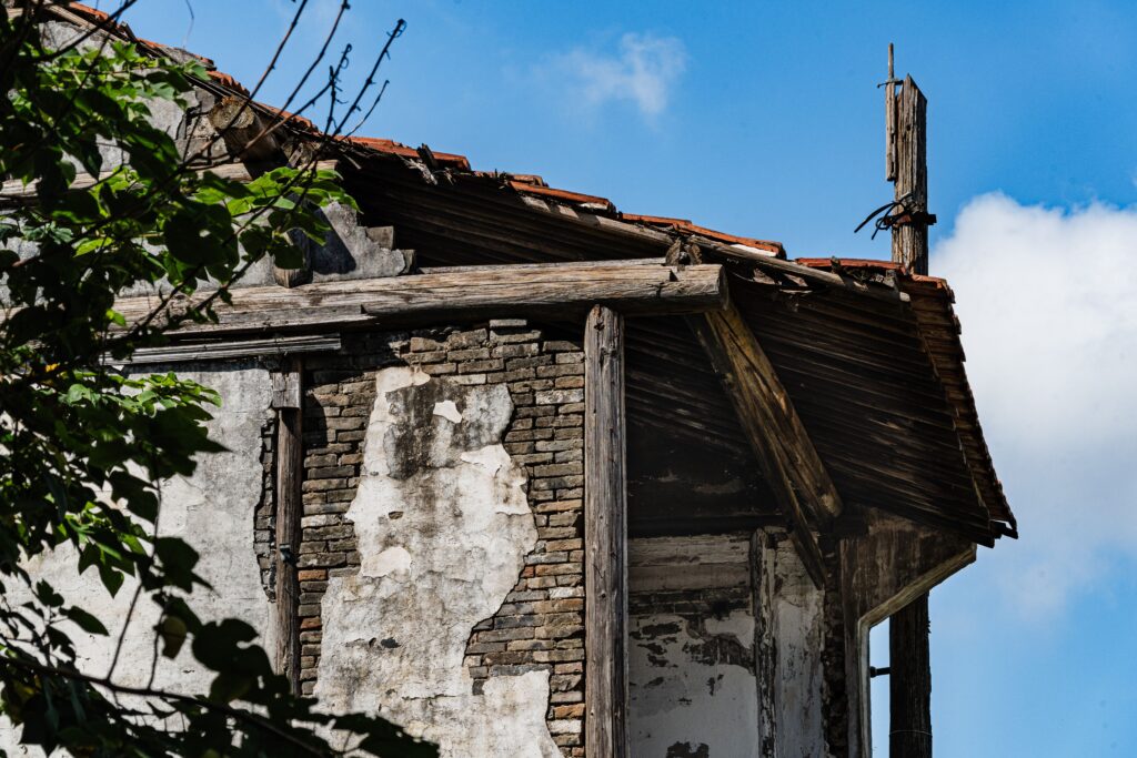 A detailed shot of an old building's exterior showing exposed bricks, crumbling plaster, and a wooden support beam holding up the edge of a damaged tiled roof.